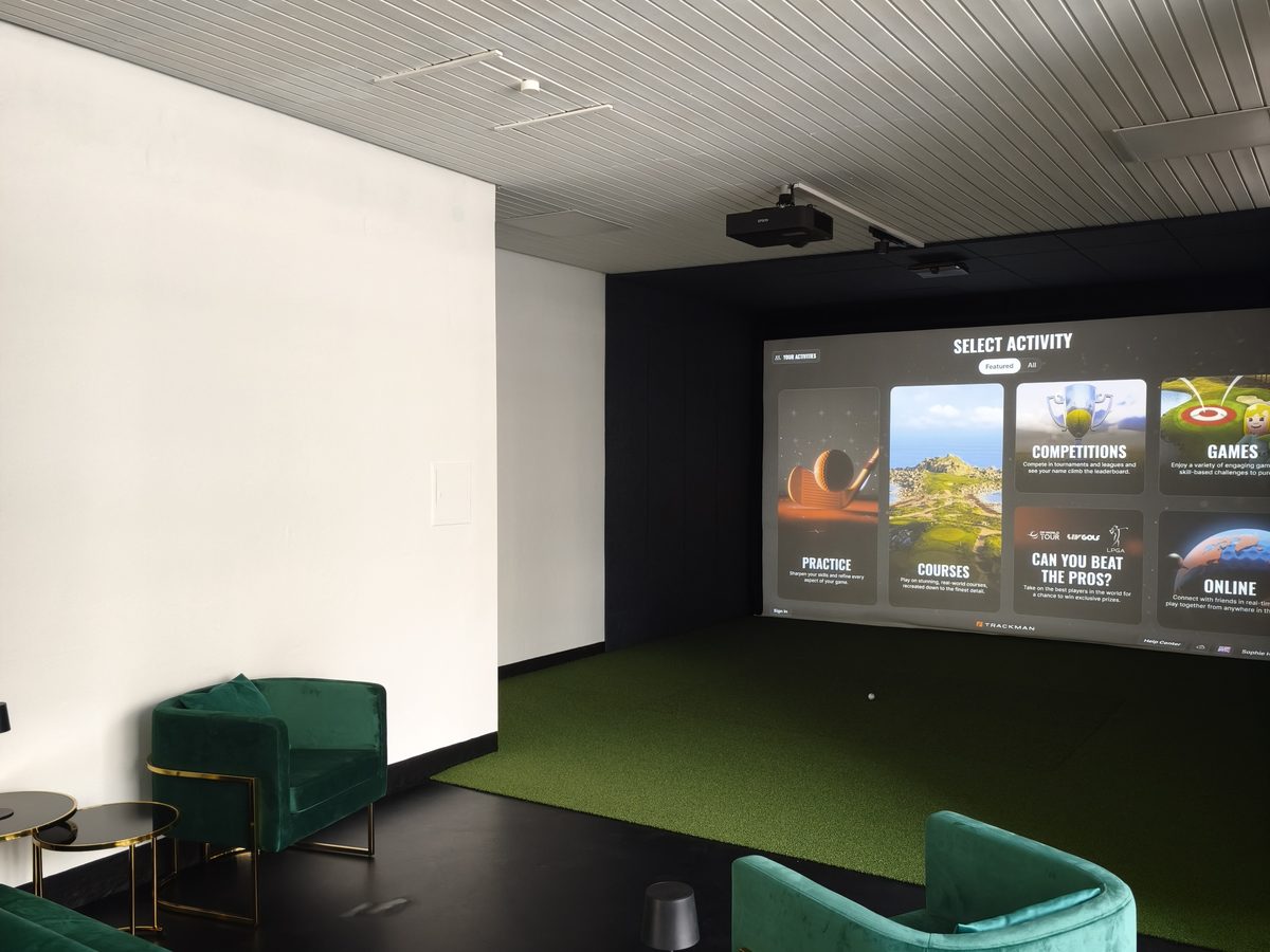 Lounge bei Hit it Hard mit grünen Samtsesseln und Blick in die Abschlagbox mit Trackman-Leinwand